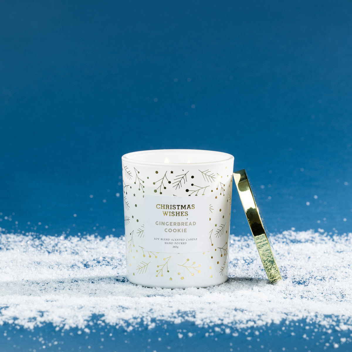 Christmas Wishes Gingerbread Cookie 283g scented candle in a white glass vessel with gold and green botanical details, lit with a gold lid resting beside it.