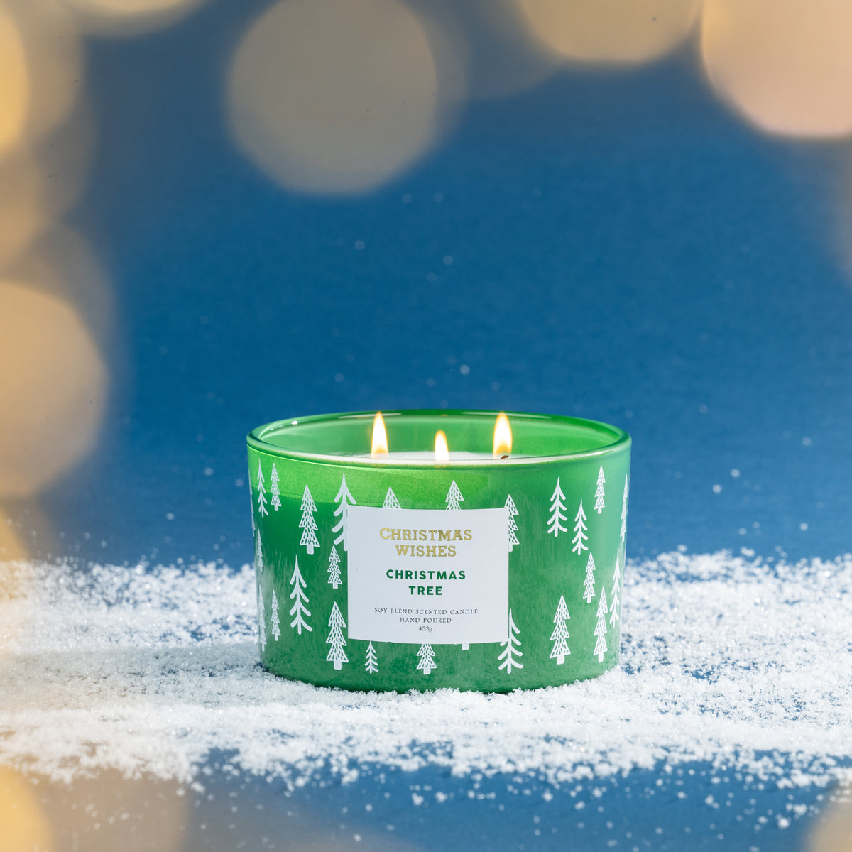 Christmas Wishes Christmas Tree 453g scented candle lit with multiple wicks against a festive blue background with warm blurred lights.