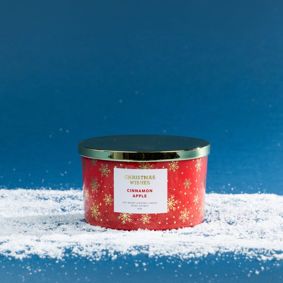 Christmas Wishes Cinnamon Apple 453g scented candle in a red vessel with gold snowflake detailing and coordinating gold lid, displayed on a snowy blue backdrop.