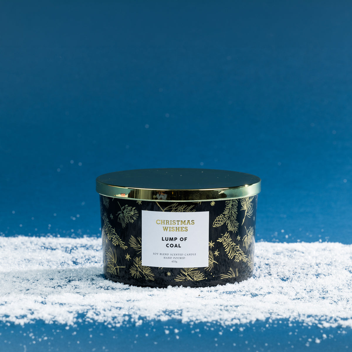 Christmas Wishes Lump of Coal 453g scented candle in a black vessel with gold botanical detailing and coordinating gold lid, displayed on faux snow.