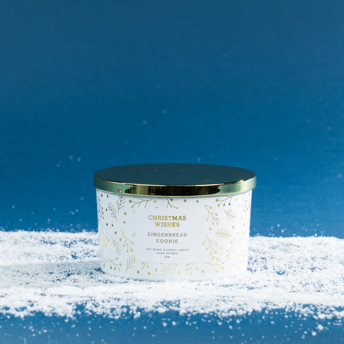 Christmas Wishes Gingerbread Cookie 453g scented candle in a white vessel with gold botanical detailing and coordinating gold lid, displayed on a snowy blue backdrop.