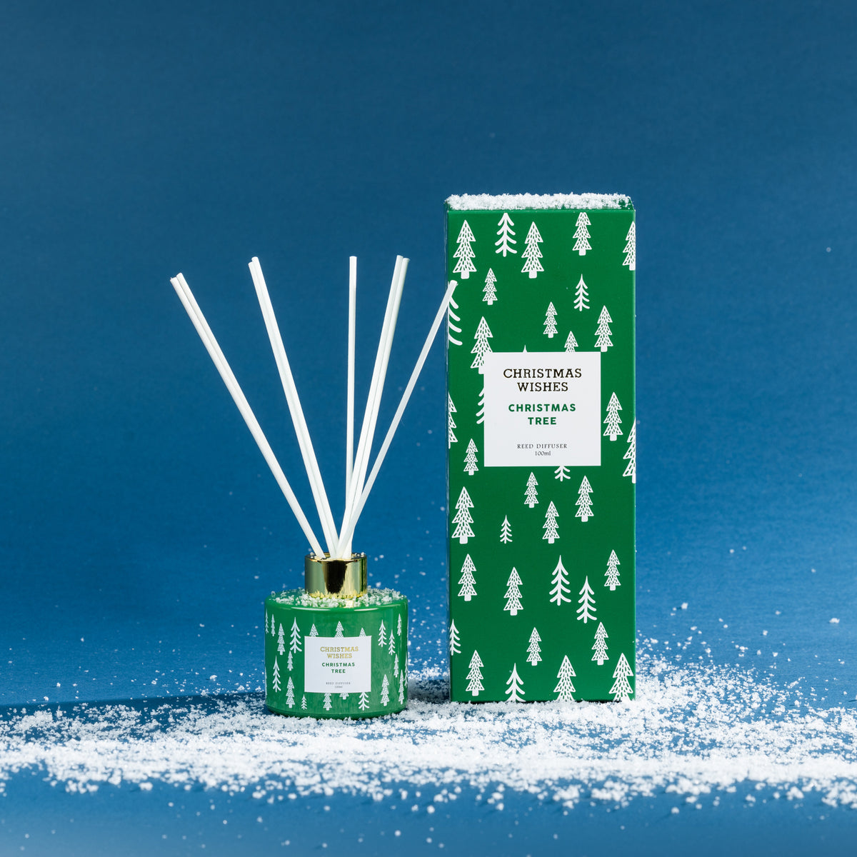 Christmas Wishes Christmas Tree 100ml reed diffuser with white reeds, gold collar and coordinating green tree-patterned gift box, displayed on a snowy blue backdrop.