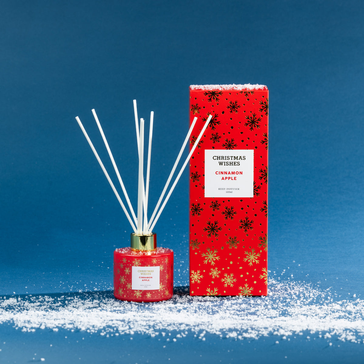 Christmas Wishes Cinnamon Apple 100ml reed diffuser with white reeds, gold collar and coordinating red snowflake-patterned gift box, displayed on a snowy blue backdrop.
