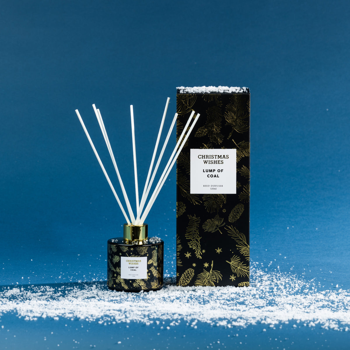 Christmas Wishes Lump of Coal 100ml reed diffuser with white reeds, gold collar and coordinating black and gold botanical gift box, displayed on a snowy blue backdrop.