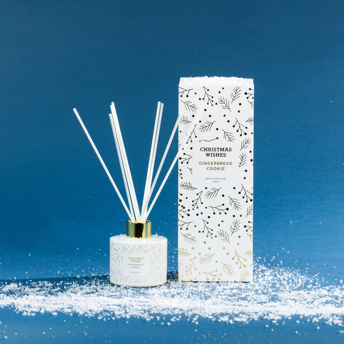Christmas Wishes Gingerbread Cookie 100ml reed diffuser with white reeds, gold collar and coordinating white botanical-patterned gift box, displayed on a snowy blue backdrop.