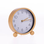 A round analog table clock with a matte gold metal frame, two small feet, and a white face with black numerals and gold hands, shown on a white background.
