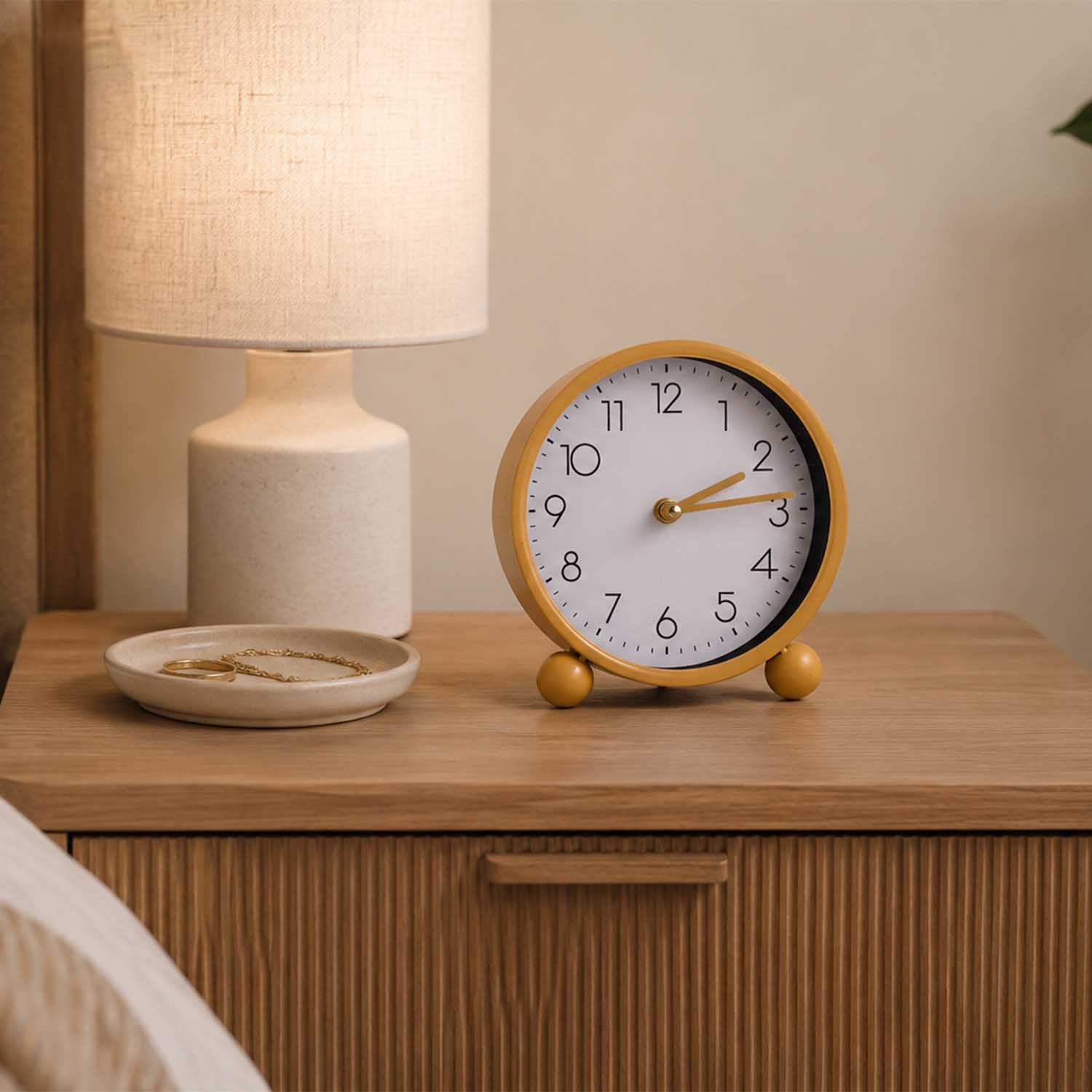 A round wooden alarm clock with a white face and gold-toned hands sits on a light wood bedside table next to a lit lamp and a small dish.
