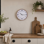 A round wall clock with a light wood frame and black numerals and hands, mounted on a beige wall above a wooden countertop with kitchen items and plants.