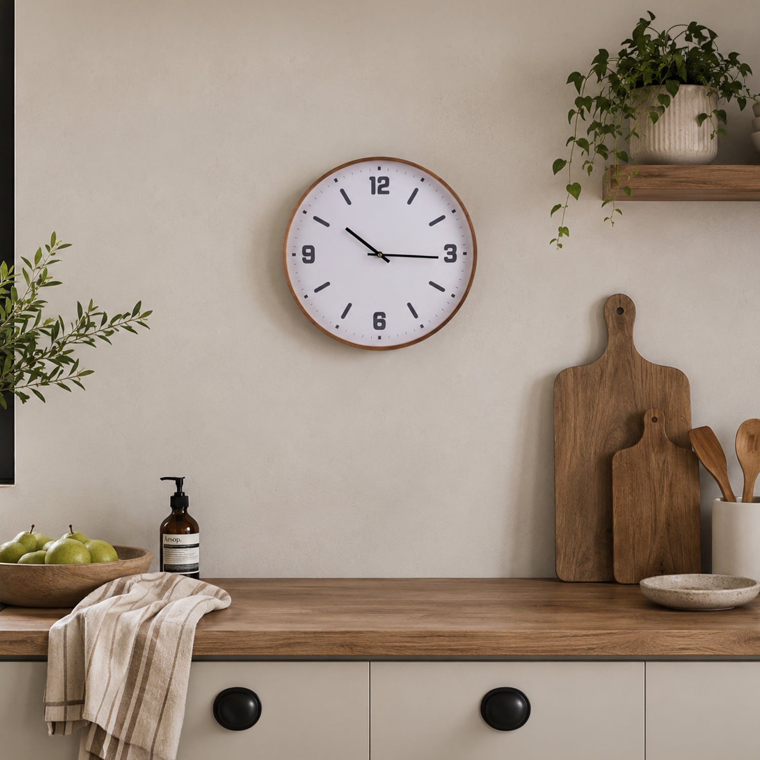 A round wall clock with a light wood frame and black numerals and hands, mounted on a beige wall above a wooden countertop with kitchen items and plants.