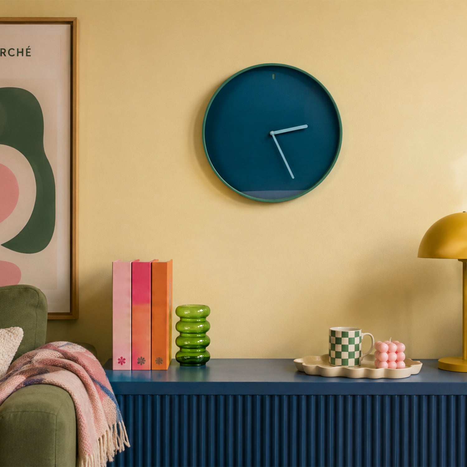 A round wall clock with a deep teal-blue face and matching outer rim, mounted on a warm beige wall above a blue ribbed sideboard. The scene includes a green armchair with a pink throw, colorful books, a green glass vase, a checkered mug, pink candles, and a gold table lamp.