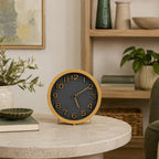 A round analog desk clock with a light wood frame and a dark charcoal face, styled on a round stone table in a cozy, modern living room setting.