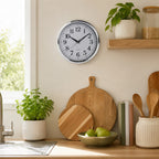 A round wall clock with a silver frame and white face hangs on a beige kitchen wall above wooden cutting boards, with potted plants, a bowl of green apples, and kitchen utensils nearby.