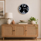A round wall clock with a black face and white numerals hangs above a light wood sideboard in a styled room. The scene includes a table lamp, small vase, books, bowl, potted plant, and framed wall art.