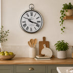 A round vintage-style wall clock with a white face, black Roman numerals, and black hands, hanging on a cream wall above a wooden countertop with kitchen items and plants.