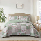 A patchwork quilted bedding set with floral and textured square patterns in sage green, dusty pink, and white, styled on a beige bed frame in a bright, modern bedroom.