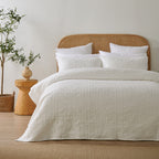 White quilted coverlet neatly styled on a bed with matching pillowcases, a woven rattan headboard and natural timber décor.
