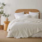 White quilted coverlet styled loosely across a bed with matching pillowcases, a woven rattan headboard and natural timber décor.
