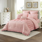 Angled view of a dusty pink comforter set with pleated panel detailing, decorative trim, ruffled edging, and matching cushions, styled in a light and elegant bedroom.