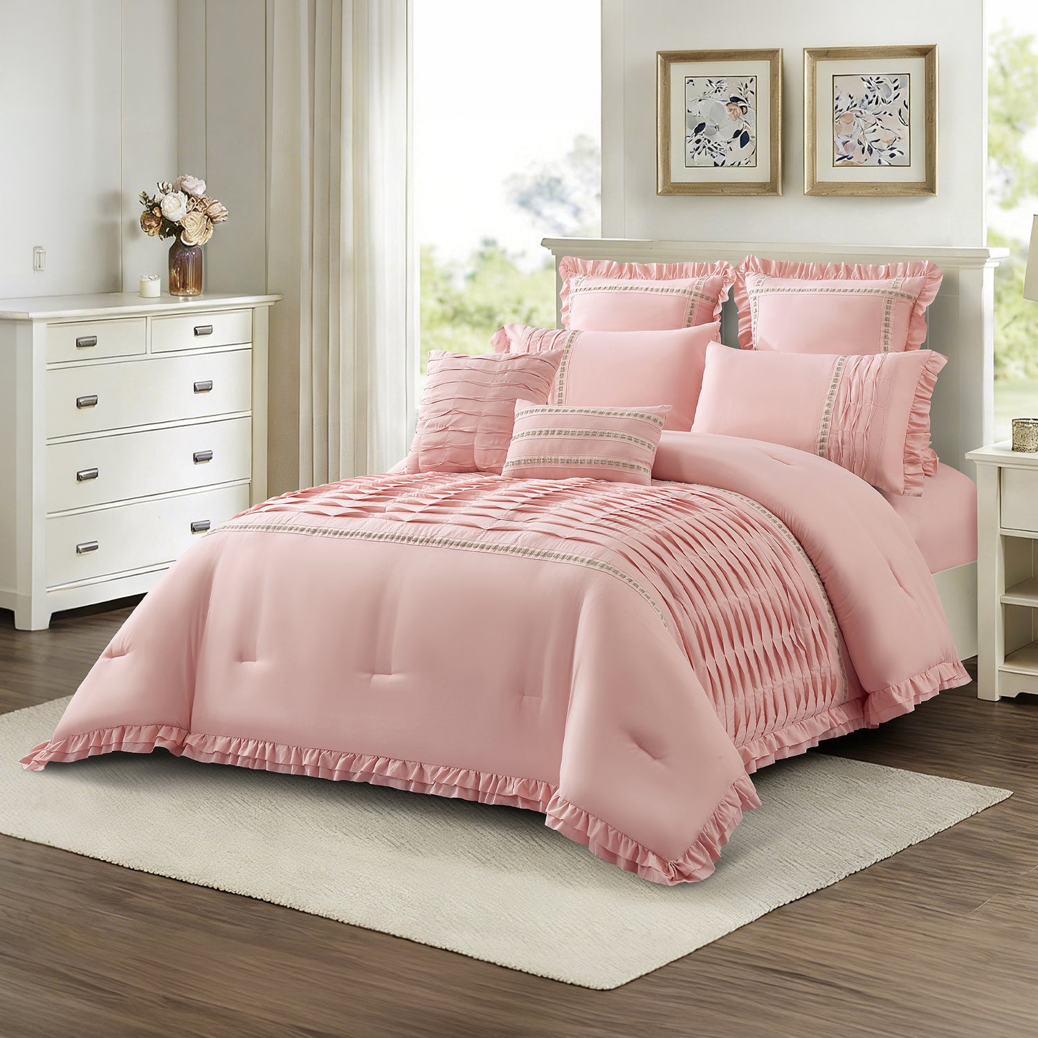 Angled view of a dusty pink comforter set with pleated panel detailing, decorative trim, ruffled edging, and matching cushions, styled in a light and elegant bedroom.