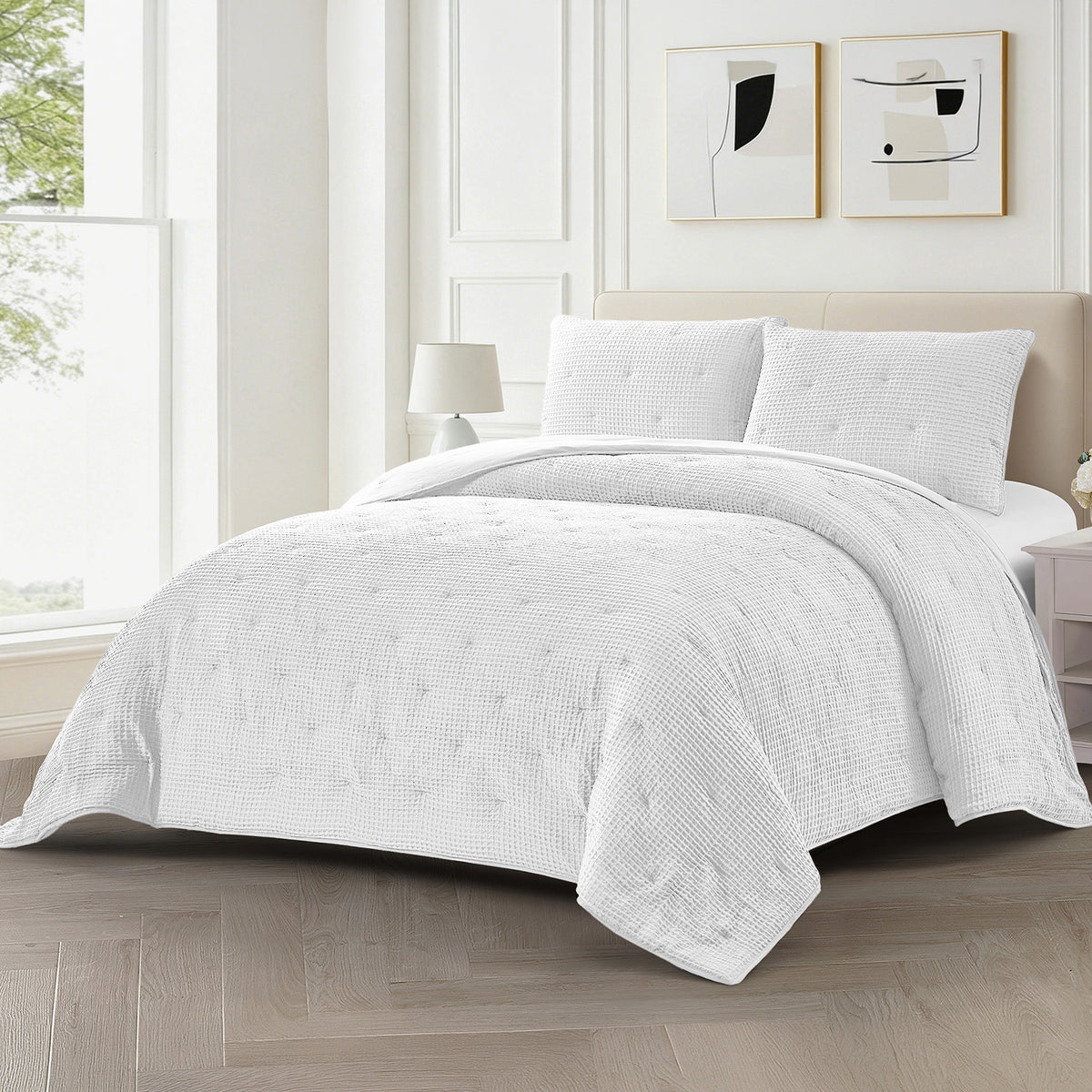 A white textured quilted comforter set with matching shams and two pillows, styled on a bed in a bright, modern bedroom with light wood flooring and neutral decor.