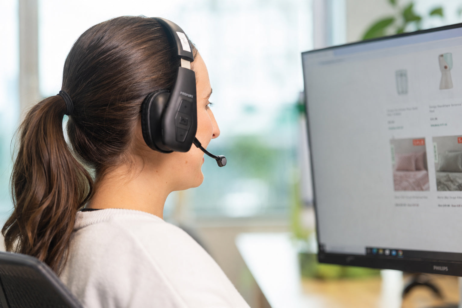 A woman in profile wearing a black over-ear headset with a microphone, seated in front of a computer monitor in a bright office setting.