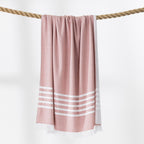 A rust and white beach towel with fringed edges hangs from a thick rope against a clean white background.