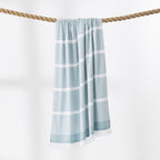 A teal and white striped beach towel with fringed edges hangs from a thick rope against a clean white background.