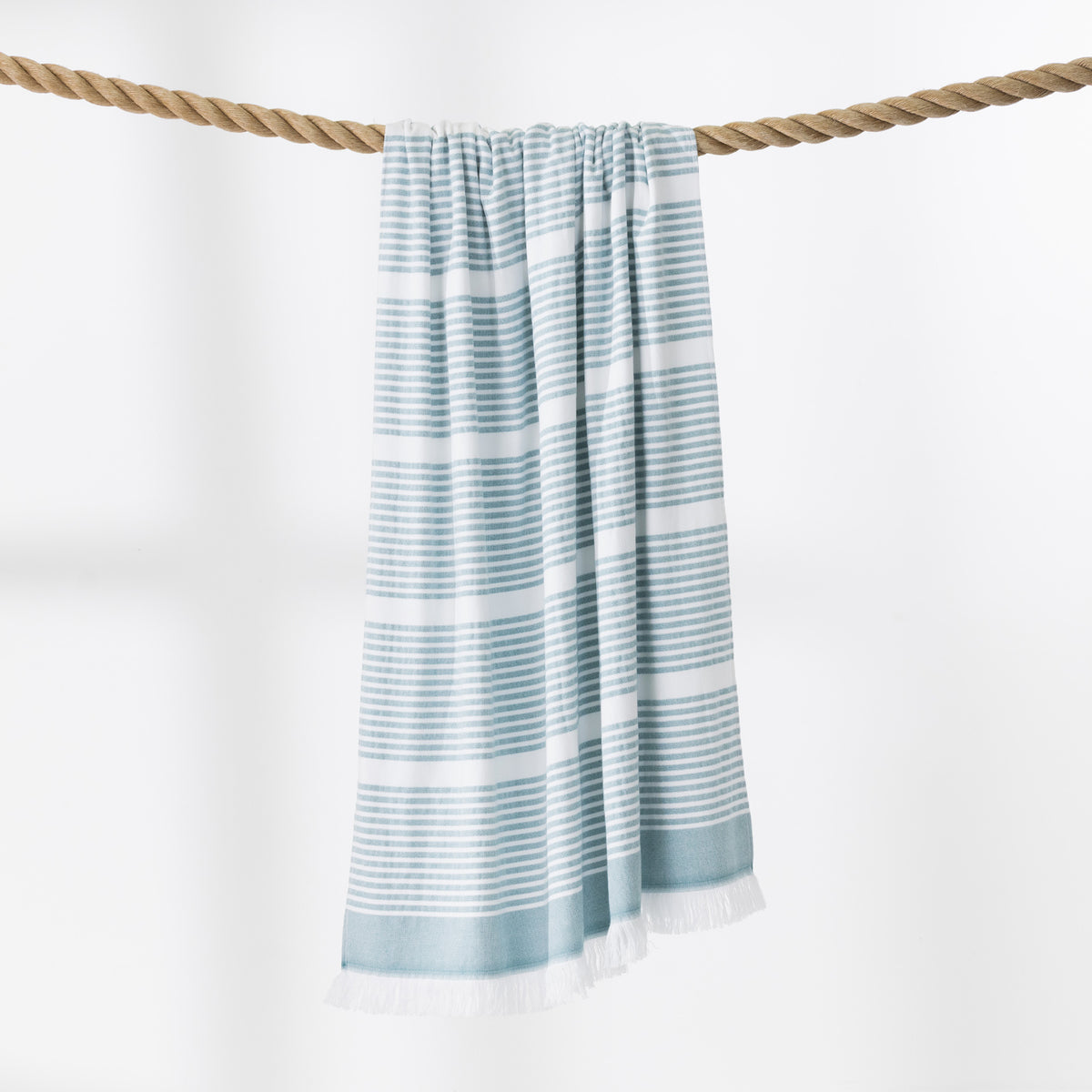 A teal and white striped beach towel with fringed edges hangs from a thick rope against a clean white background.