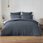Blue textured bedding set on a bed in a bedroom with wooden flooring and a striped wall.