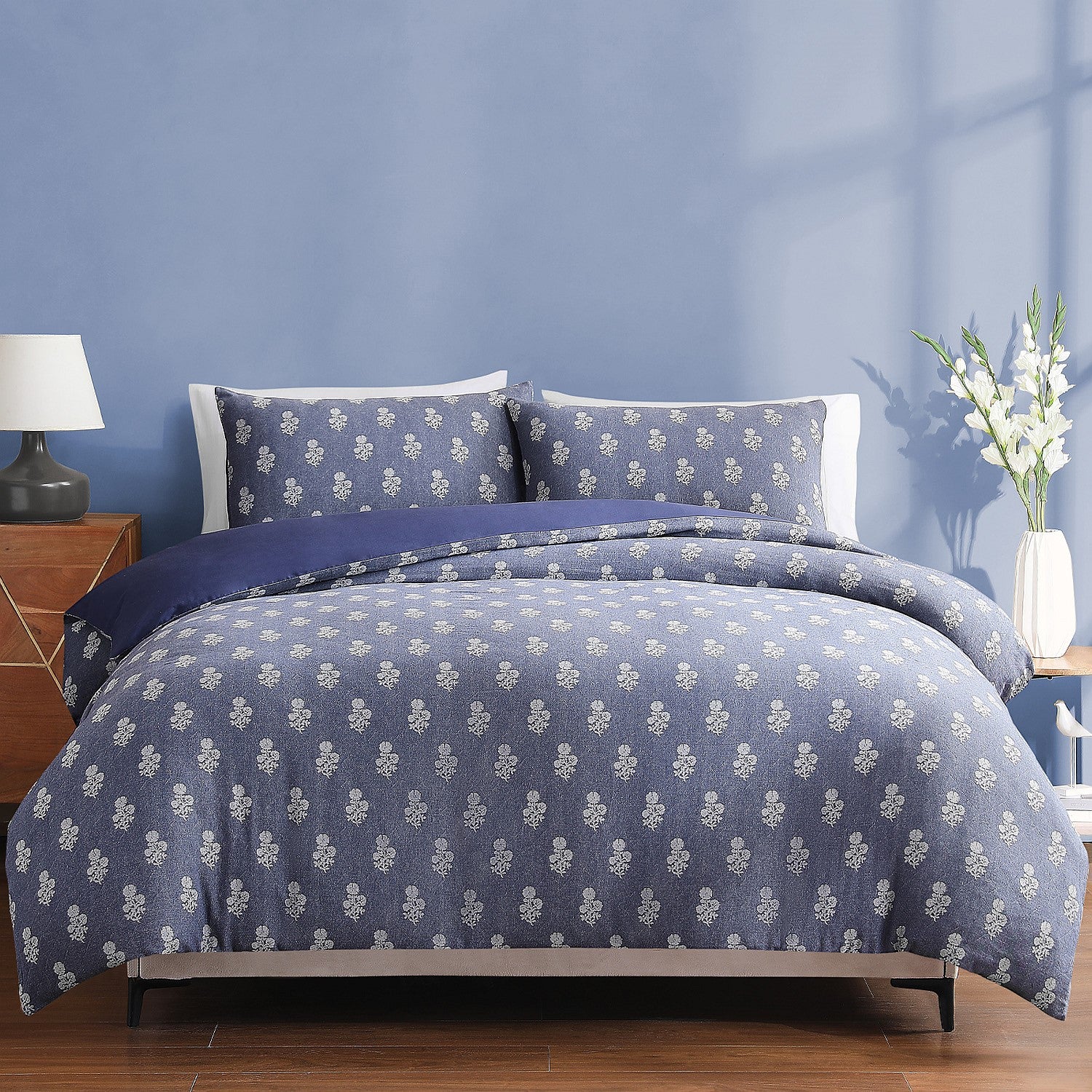 Blue floral patterned bedding set on a bed with a blue wall and wooden nightstand.
