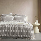 A neatly made bed dressed in a gray and white geometric checkered bedding set, styled in a minimalist bedroom with a textured beige wall, a small cream-colored side table, and a vase.