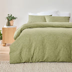 Green bedding set on a bed with a wooden nightstand and plants in the background.