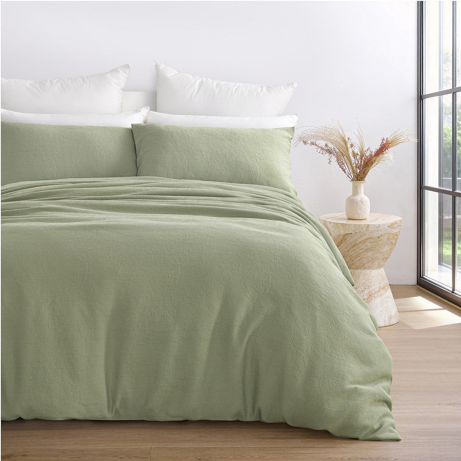 Green bedding set on a bed in a bright room with a vase on a side table.