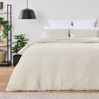 Beige comforter on a bed in a bedroom with plants and shelves.