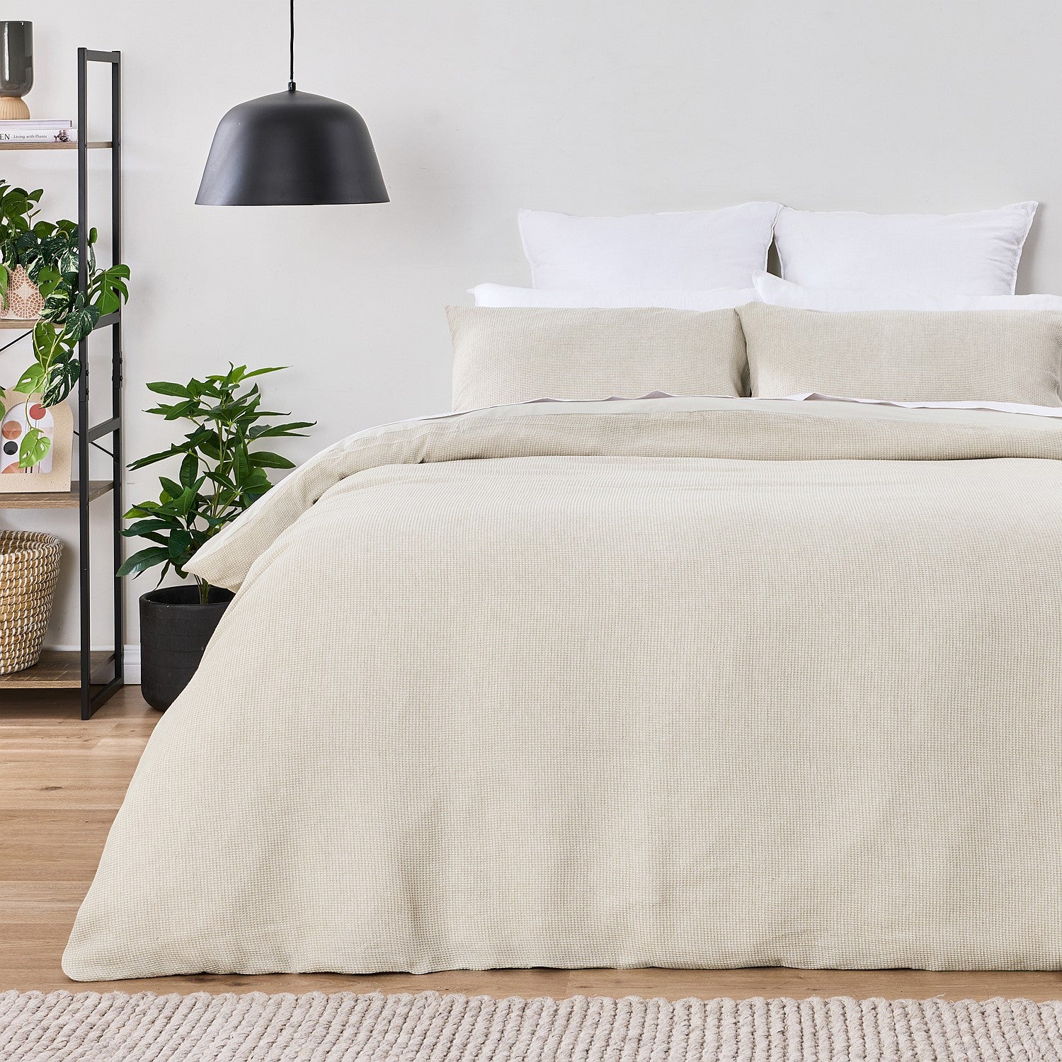 Beige comforter on a bed in a bedroom with plants and shelves.