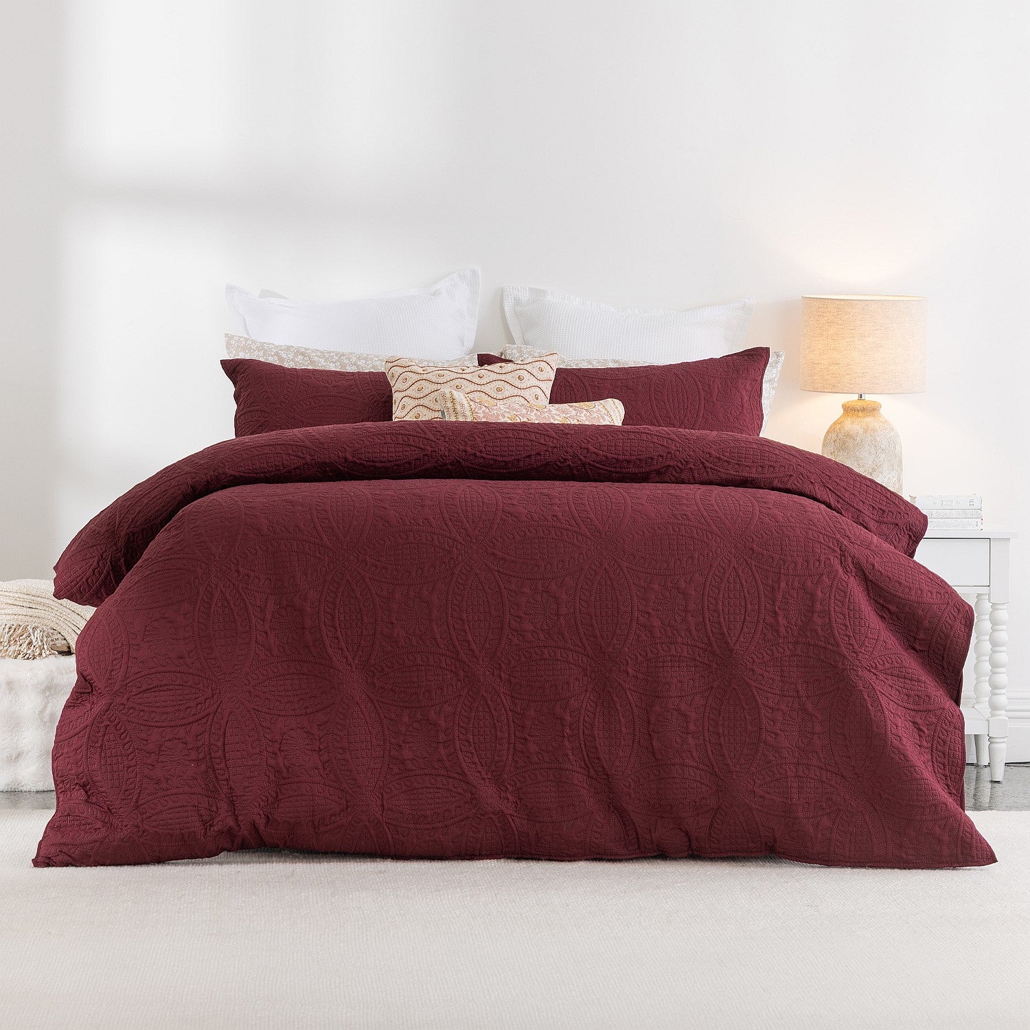 Burgundy quilted coverlet on a bed with pillows and a lamp in the background.