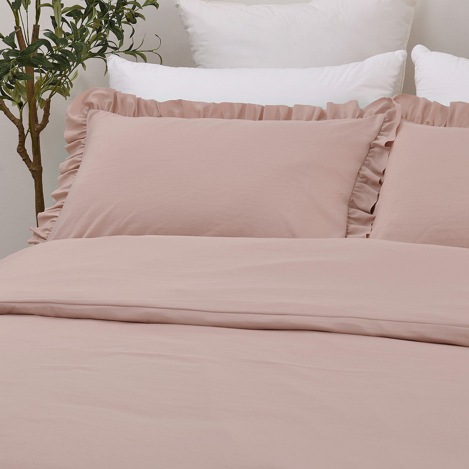 Pink bedding set with ruffled edges on a bed, featuring white pillows and a plant in the background.