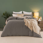 A styled bed dressed in a gray bedding set with white pillows, a beige patterned accent pillow, and a cream fringed throw blanket, set against a dark green wall with a lit bedside lamp.