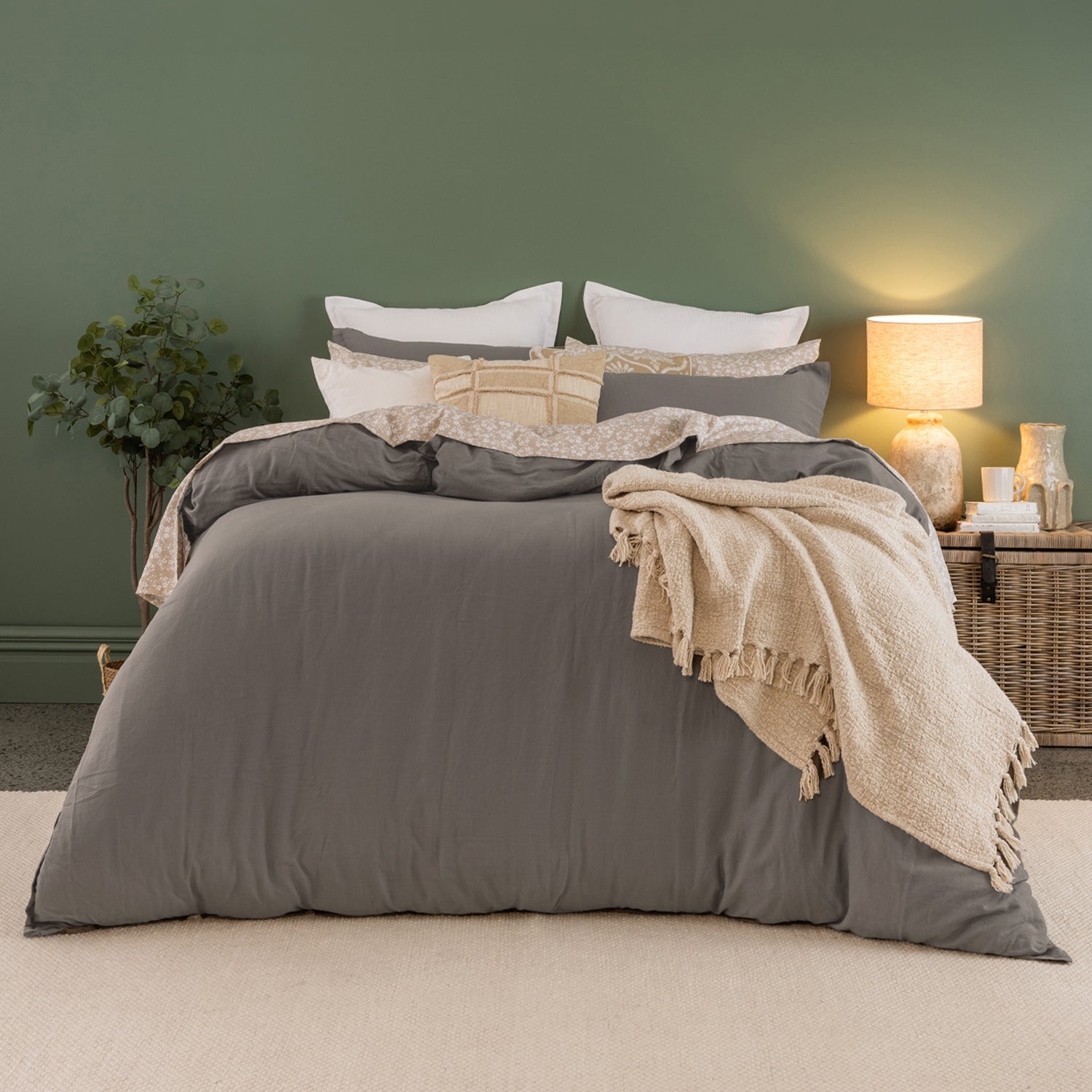 A styled bed dressed in a gray bedding set with white pillows, a beige patterned accent pillow, and a cream fringed throw blanket, set against a dark green wall with a lit bedside lamp.