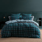 A bed dressed in a teal and dark blue geometric patterned duvet cover and matching pillow shams, styled in a modern bedroom with a dark green paneled wall and a woven beige rug.