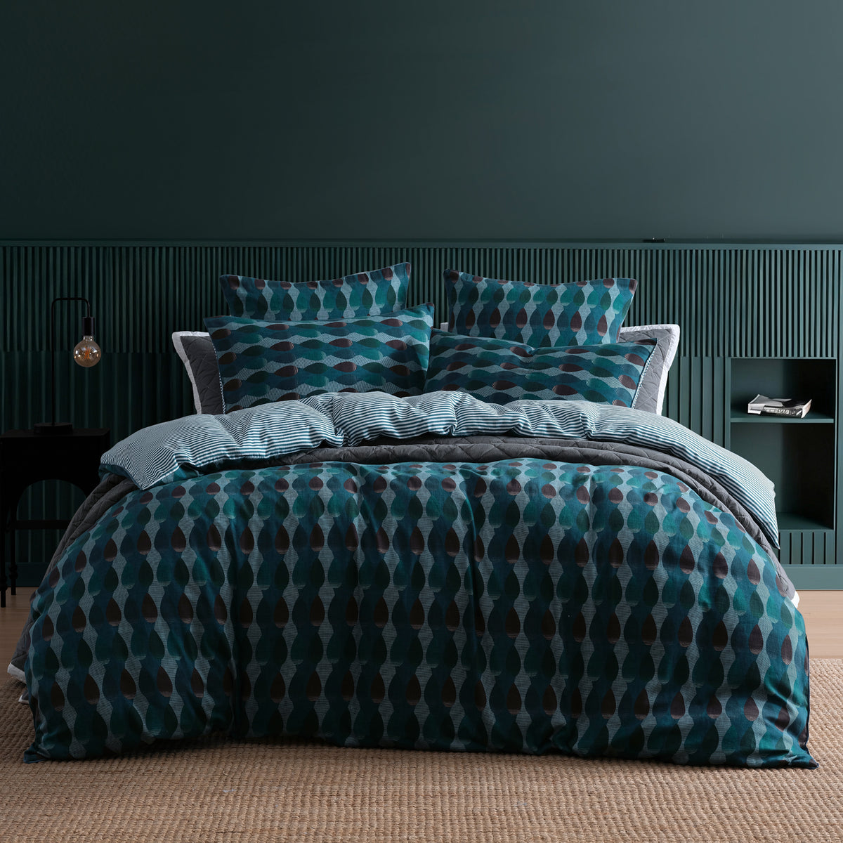 A bed dressed in a teal and dark blue geometric patterned duvet cover and matching pillow shams, styled in a modern bedroom with a dark green paneled wall and a woven beige rug.
