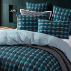 A styled bedding set on a bed in a modern bedroom, featuring a duvet cover and matching pillowcases with a teal, blue, and brown geometric pattern, layered with a light blue striped duvet and dark gray quilted sheets.