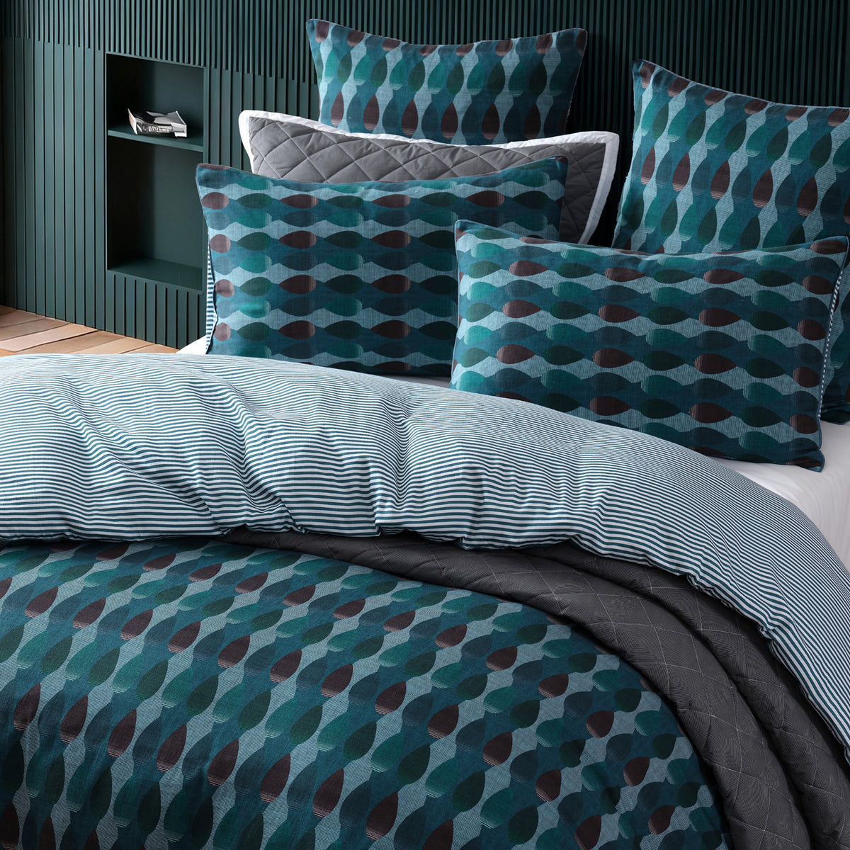 A styled bedding set on a bed in a modern bedroom, featuring a duvet cover and matching pillowcases with a teal, blue, and brown geometric pattern, layered with a light blue striped duvet and dark gray quilted sheets.