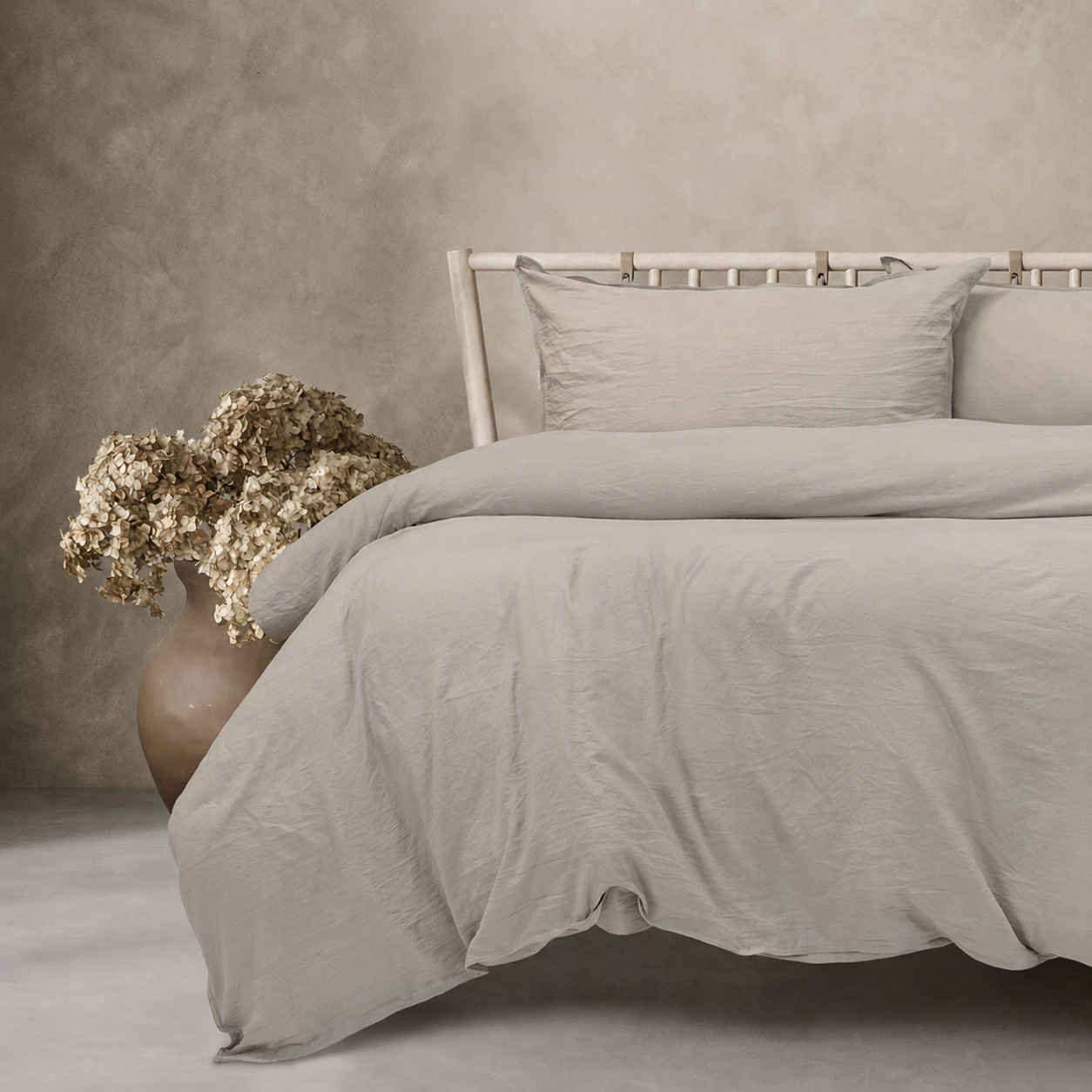 Neatly made bed with beige bedding and pillows, accompanied by a vase with dried flowers on a neutral background.