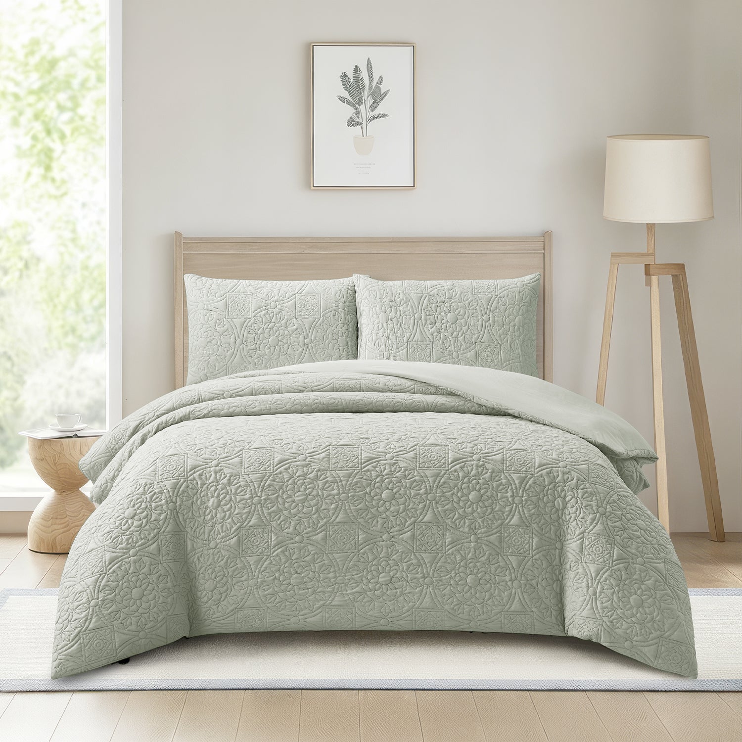 A light sage green quilted bedding set with matching pillowcases on a bed in a bright, modern bedroom. The room features a light wood headboard, a white tripod floor lamp, a small wooden side table, and a framed botanical print on the wall.