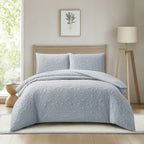 A light blue quilted bedding set with two matching pillowcases on a bed in a modern, sunlit bedroom. The room features a light wood floor, a beige wall with a framed botanical print, and a wooden tripod floor lamp.