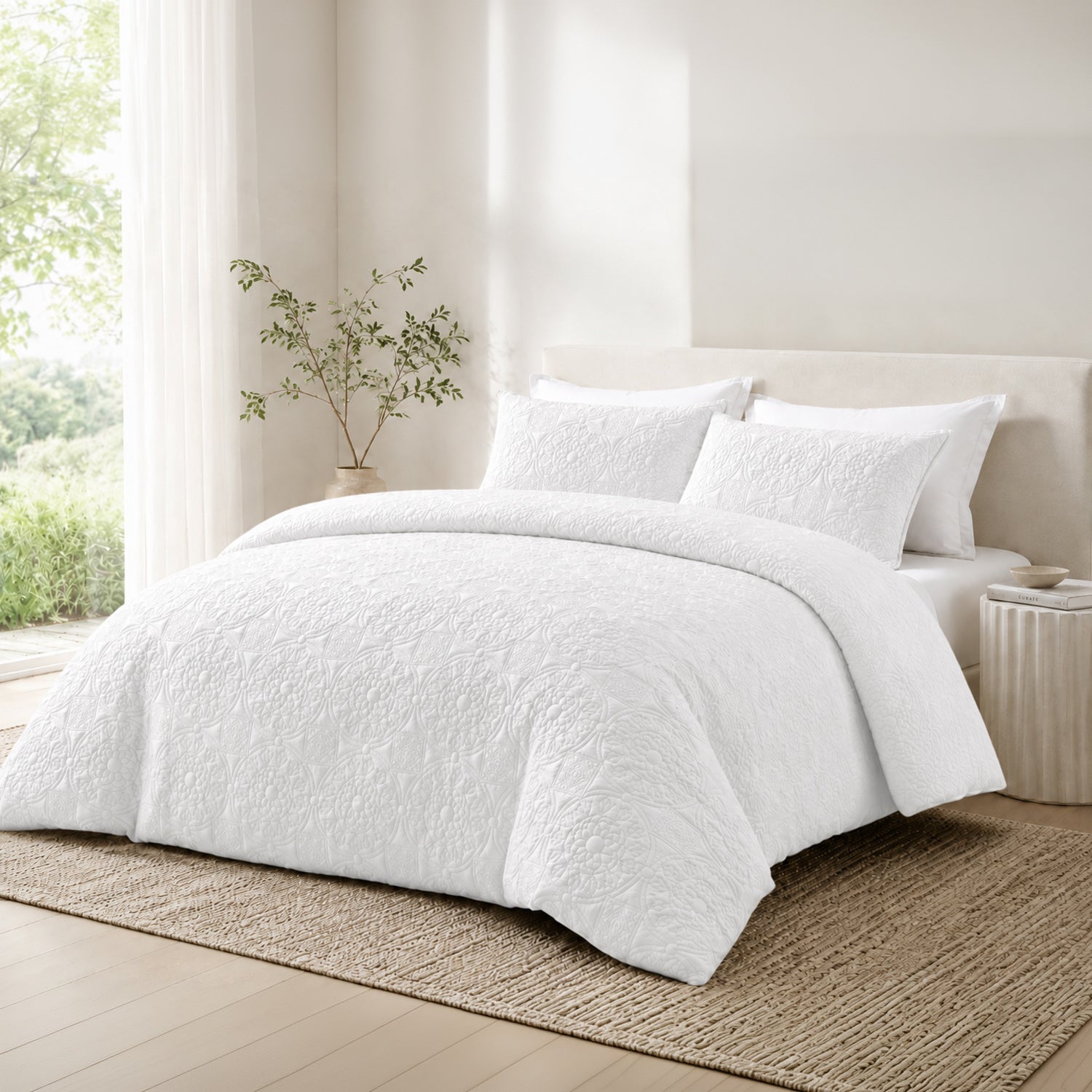 A neatly made bed in a bright, neutral bedroom setting, dressed with a textured white duvet cover and matching pillow shams. The bed is styled with a beige upholstered headboard, a woven jute rug, and a small side table with books, with soft natural light coming from a window on the left.
