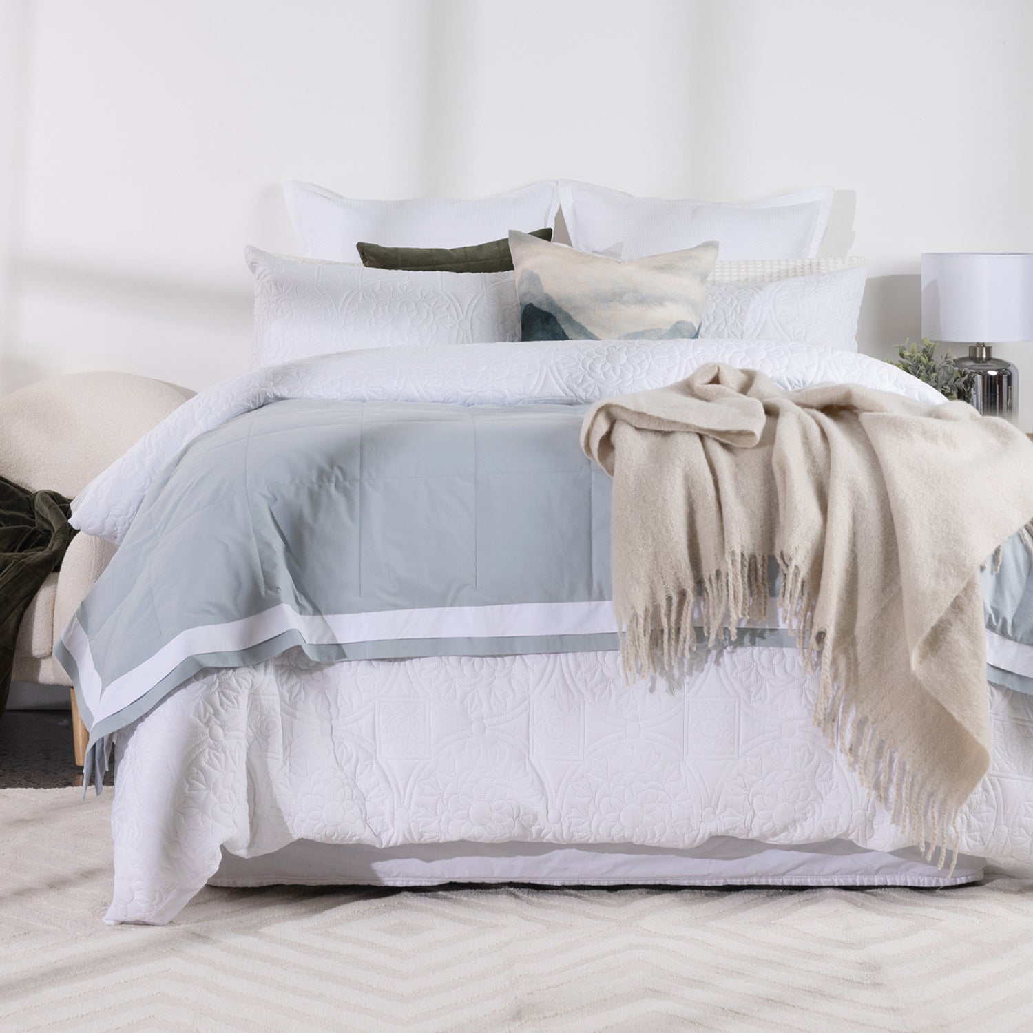 A neatly made bed styled for bedding, featuring a white quilted duvet cover set with a light blue blanket, white pillows, a beige throw blanket draped over the side, and a small accent pillow, set in a bright, modern bedroom.