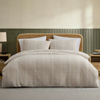 A neatly made bed in a styled bedroom setting, featuring a textured beige and white striped duvet cover and matching pillow shams. The bed has a natural rattan headboard, flanked by matching nightstands with a lamp and books, set against a beige wall with green wainscoting.