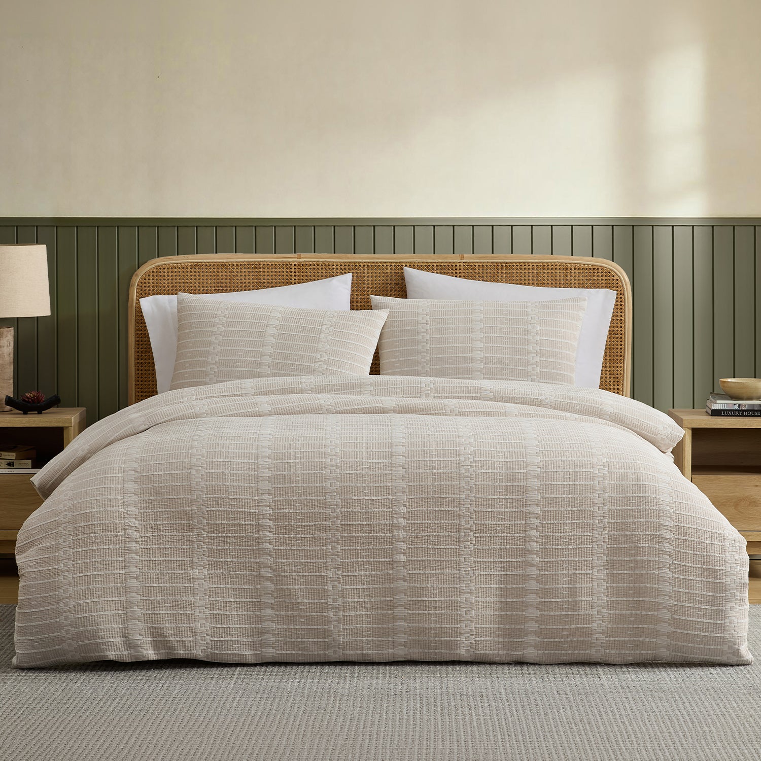 A neatly made bed in a styled bedroom setting, featuring a textured beige and white striped duvet cover and matching pillow shams. The bed has a natural rattan headboard, flanked by matching nightstands with a lamp and books, set against a beige wall with green wainscoting.